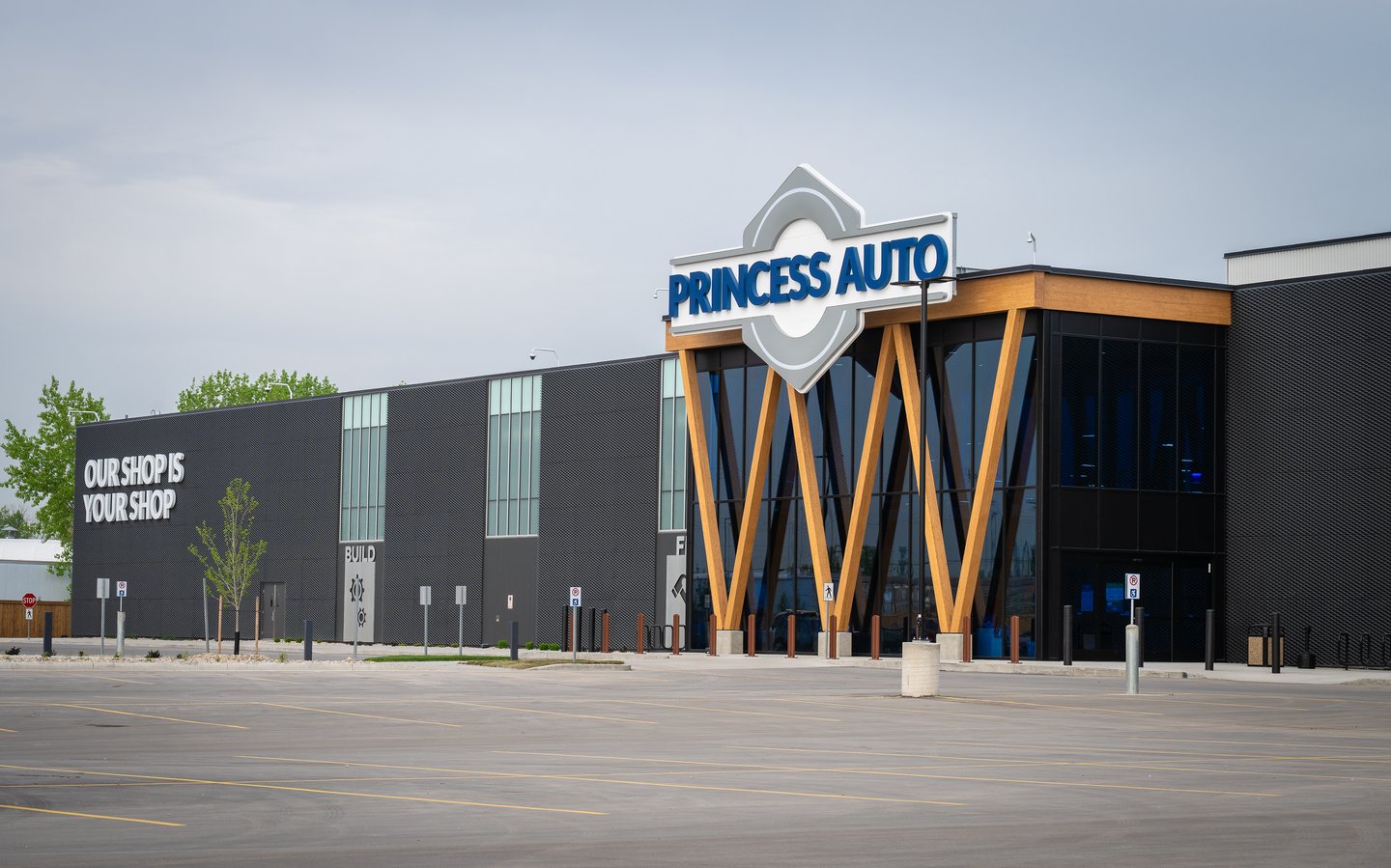 The Princess Auto flagship exterior from the parking lot, with 'Our shop is your shop' on the dark facade and the glass entrance