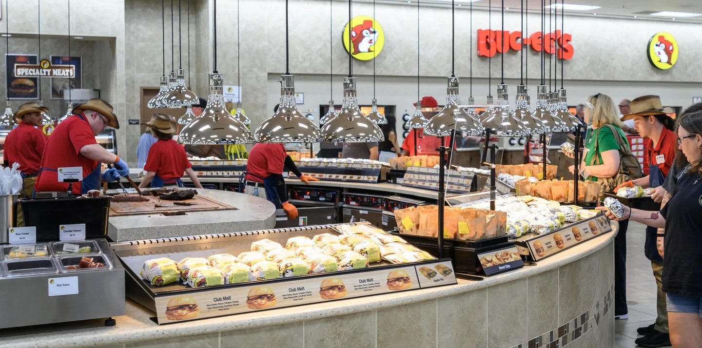 Staff in red shirts serving at the Buc-ee's food counter, with cases of prepared food under heat lamps