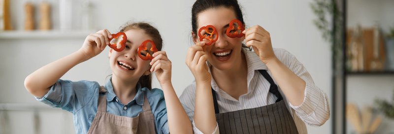A mother and daughter in aprons laughing with pepper rings held over their eyes in a kitchen