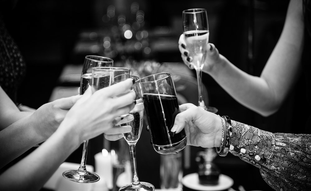 A black and white photo of friends clinking glasses of wine and beer over a dinner table