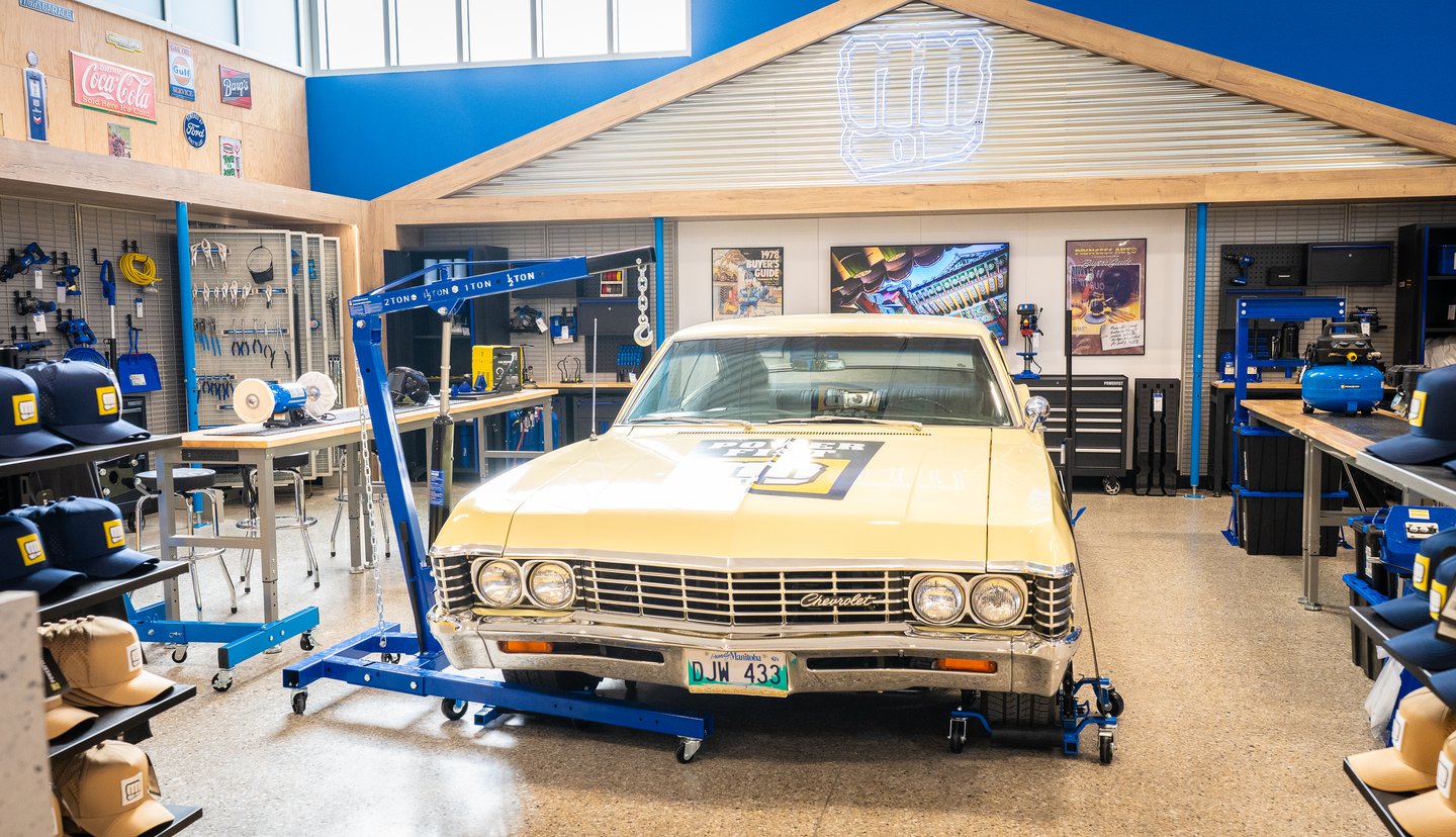 A yellow 1967 Chevelle on a lift inside the Powerfist garage display, surrounded by tools and workbenches