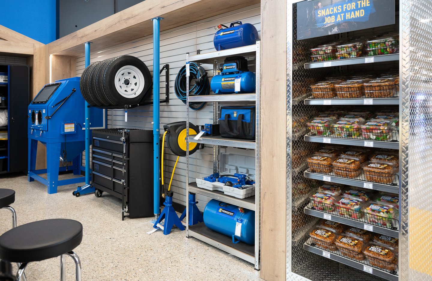 Powerfist tools on garage shelving: compressors, a sandblast cabinet, tires and a tool chest, beside a 'Snacks for the job at hand' cooler