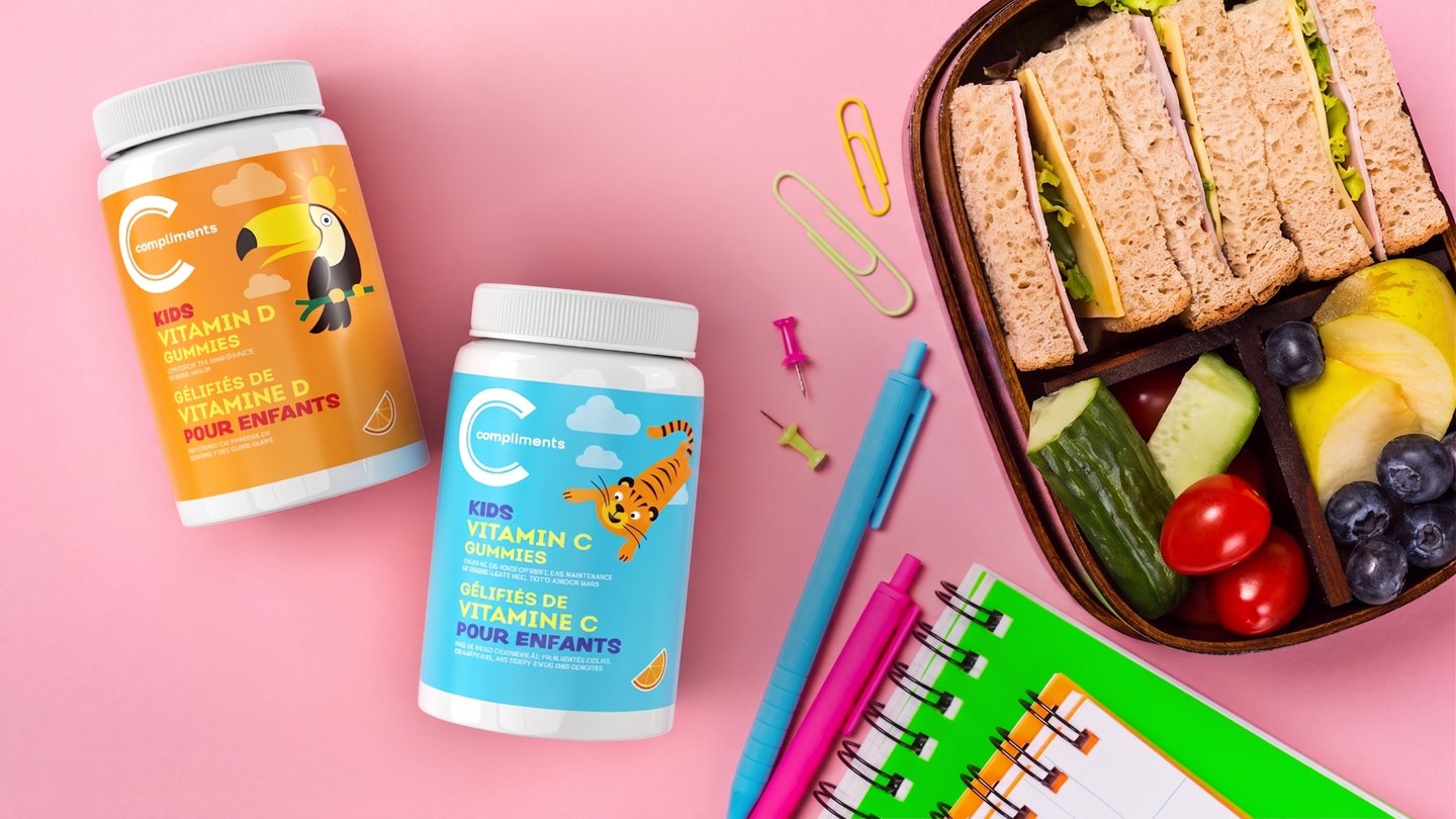 Compliments Kids Vitamin D and Vitamin C gummies on a pink desk with a packed lunch box and school supplies