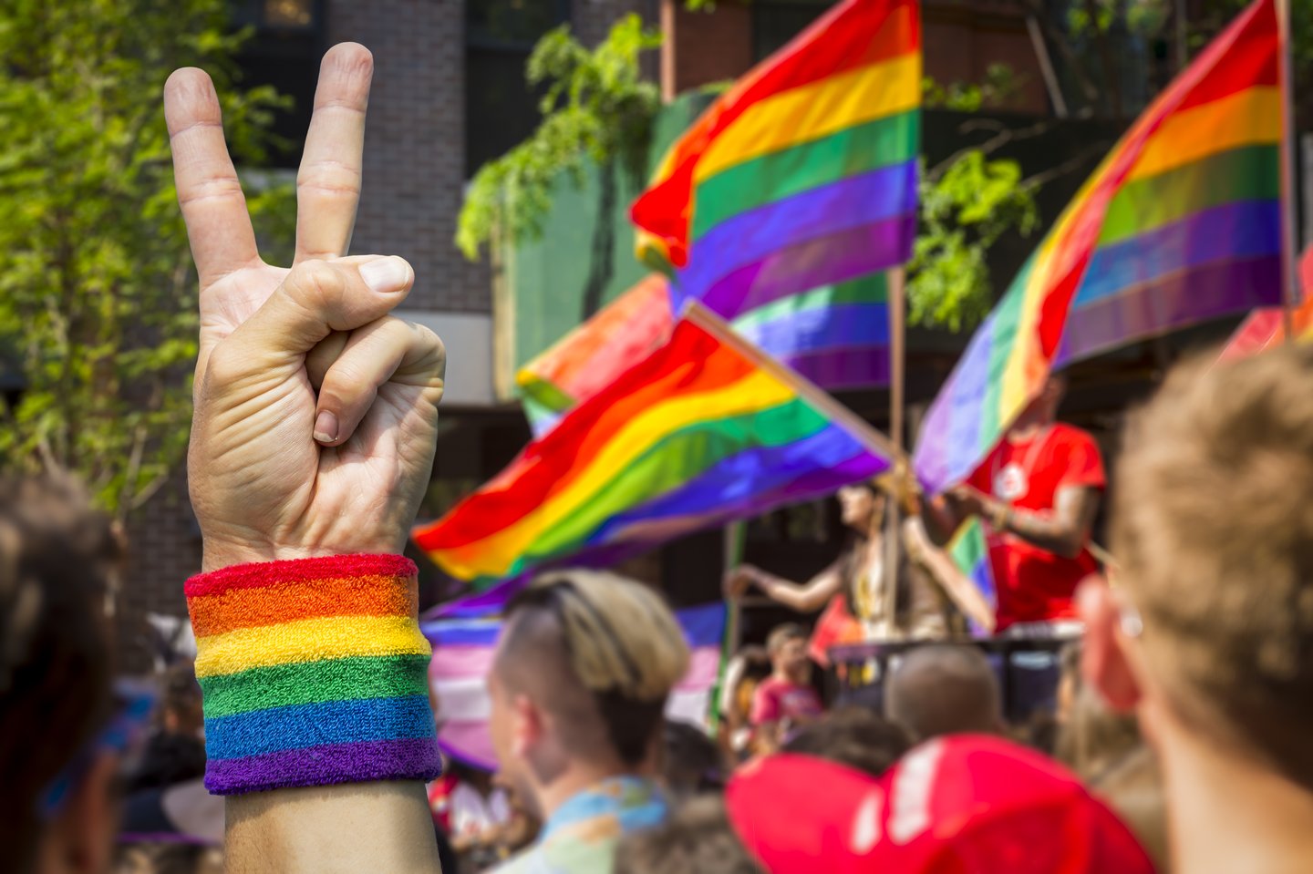 A hand making a peace sign, wearing a rainbow wristband, in front of rainbow flags at a Pride parade
