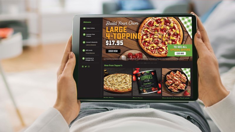 A woman holds a tablet showing the Topper's Pizza online ordering page, with a 'Build your own large 4-topping' offer