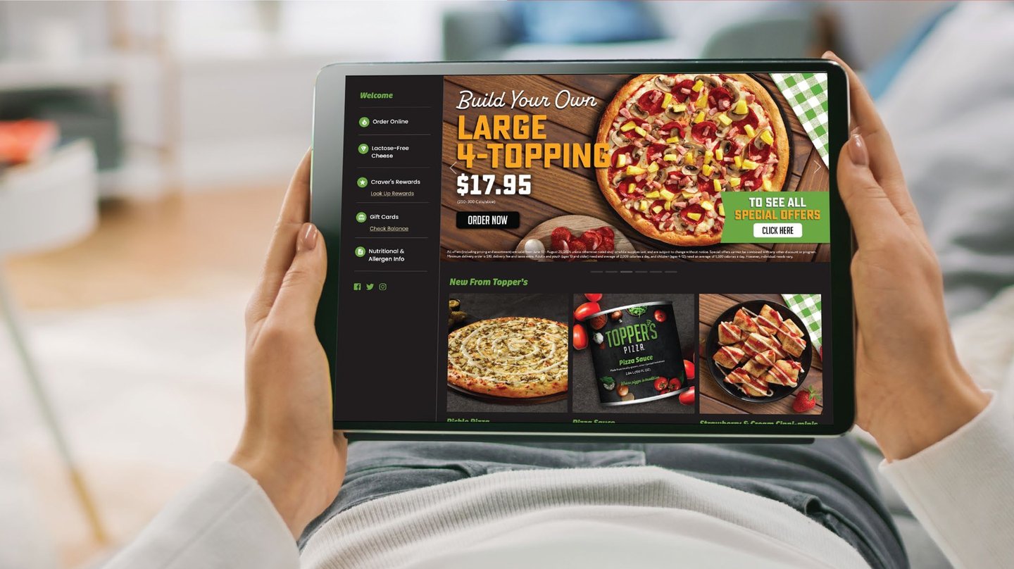 A woman holds a tablet showing the Topper's Pizza online ordering page, with a 'Build your own large 4-topping' offer