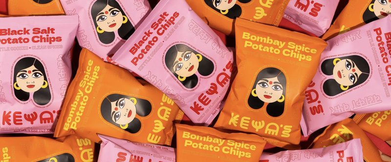 A pile of Keya's Black Salt and Bombay Spice potato chip bags in pink and orange, each with an illustrated woman's face