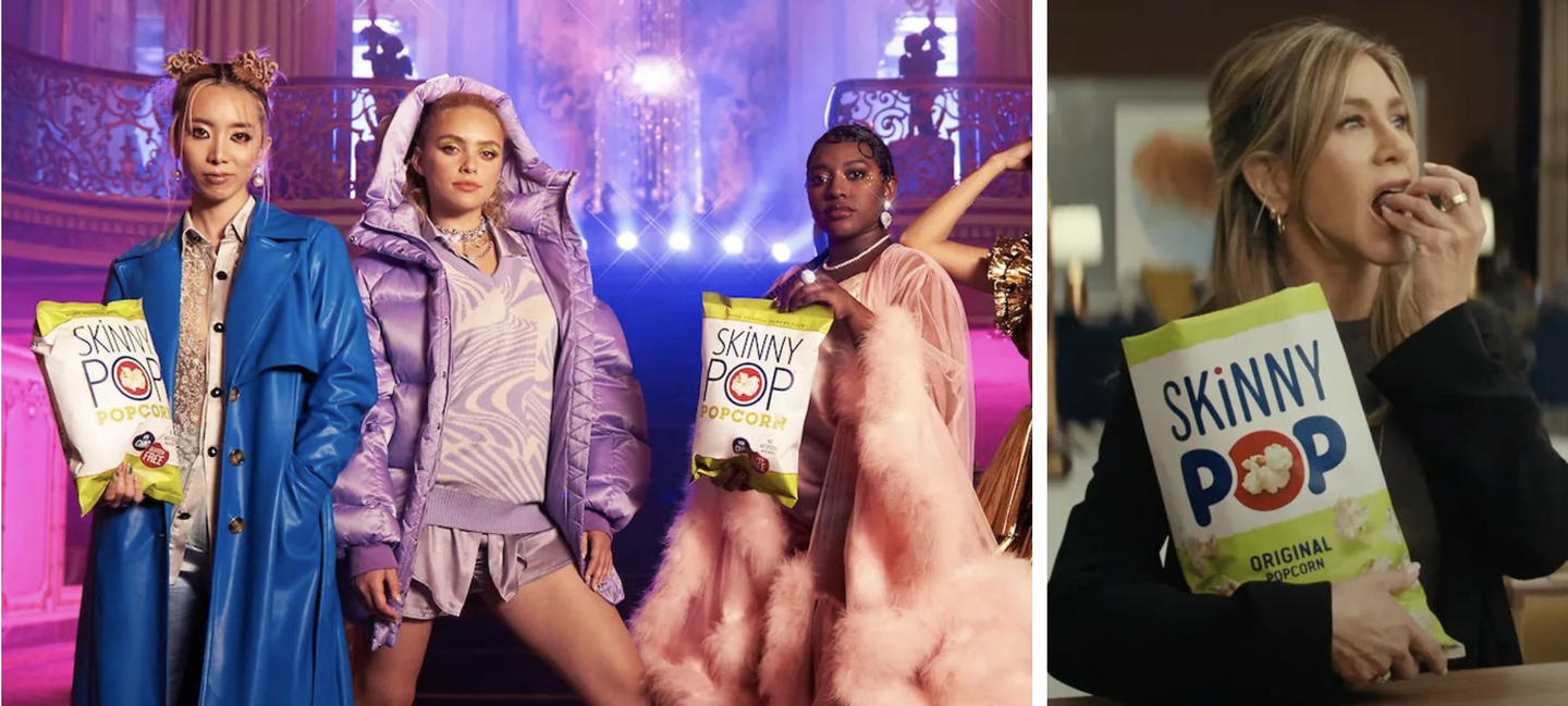 Three women in colourful coats holding SkinnyPop bags on a purple stage set, beside Jennifer Aniston eating SkinnyPop