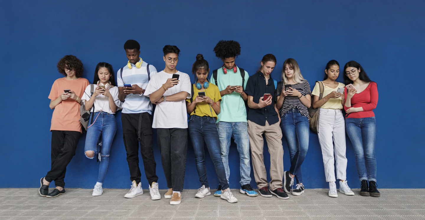 A row of teenagers lined up against a blue wall, each looking down at their phone