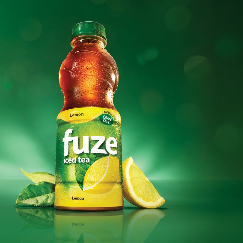 A bottle of Fuze Lemon iced tea with a lemon wedge and tea leaves, lit against a dark green background
