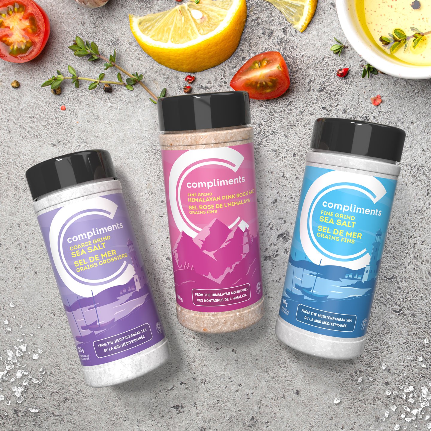 Three Compliments sea salt shakers in purple, pink and blue on a stone counter with tomatoes and lemon
