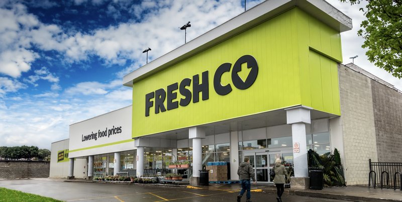 The FreshCo storefront under a blue sky, with the lime green fascia sign and 'Lowering food prices' tagline