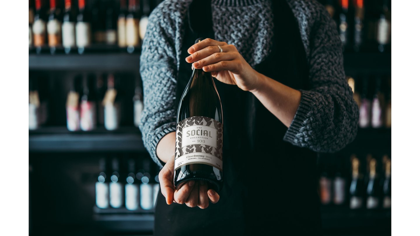 A sommelier in a wine shop holds a bottle of The Social Collection wine toward the camera