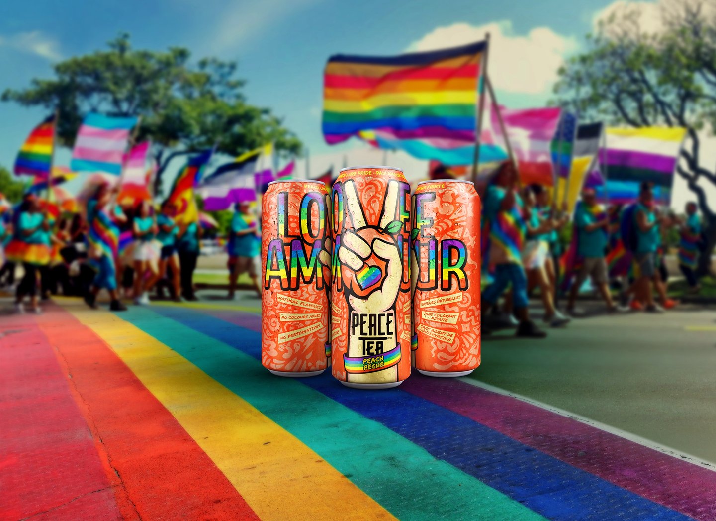 Three Peace Tea cans spelling 'Love' and 'Amour' with peace-sign hands, standing on a rainbow crosswalk at a Pride parade