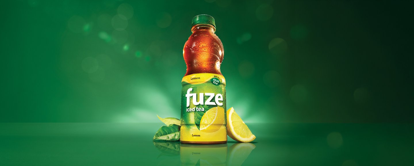 A bottle of Fuze Lemon iced tea with a lemon wedge and tea leaves, lit against a dark green background