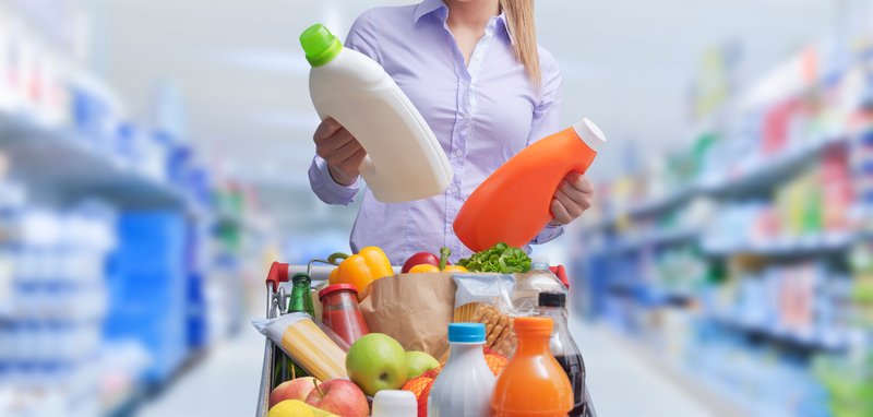 A shopper comparing two bottles in a grocery aisle, with a cart of groceries in front of her
