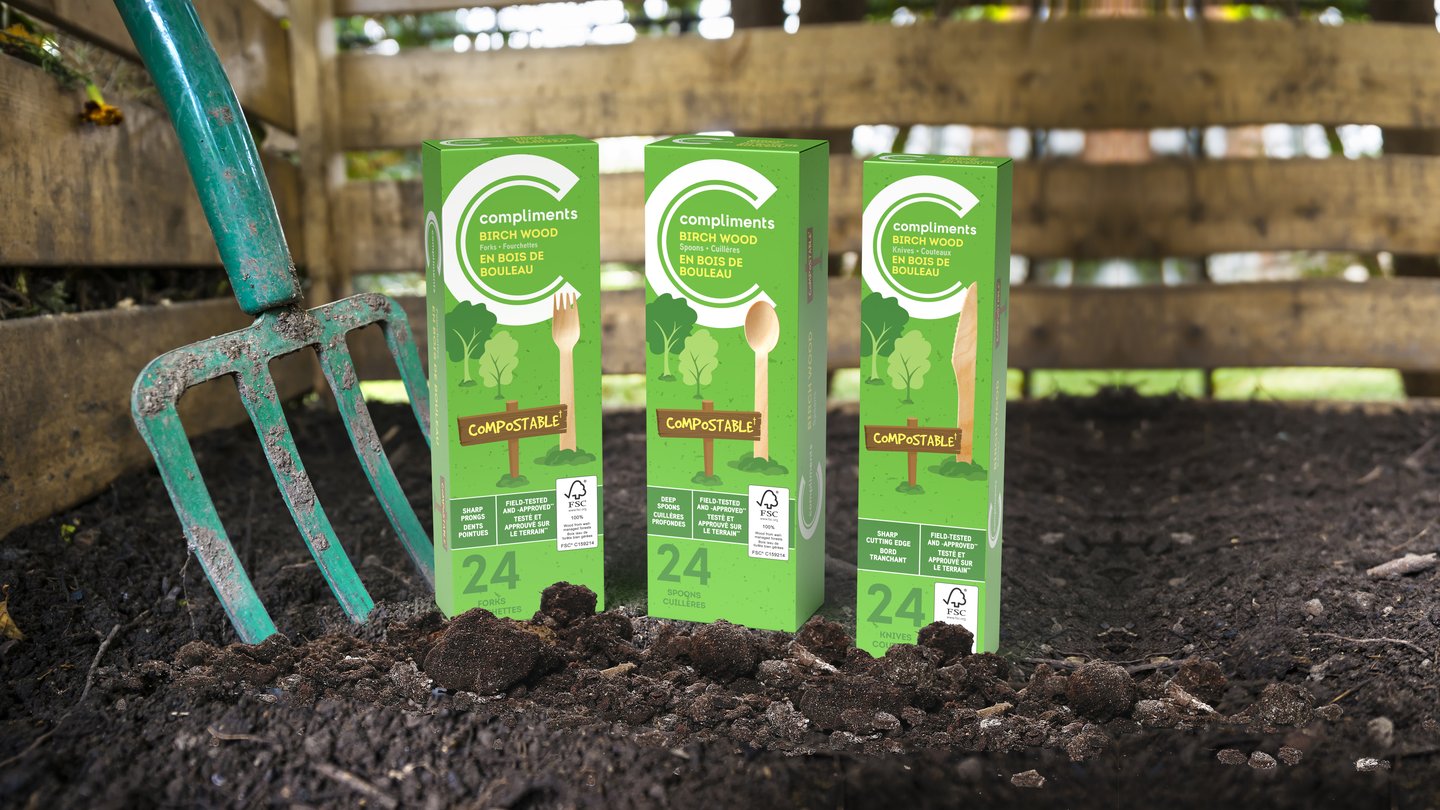 Three boxes of Compliments compostable birch wood cutlery standing in garden soil beside a garden fork