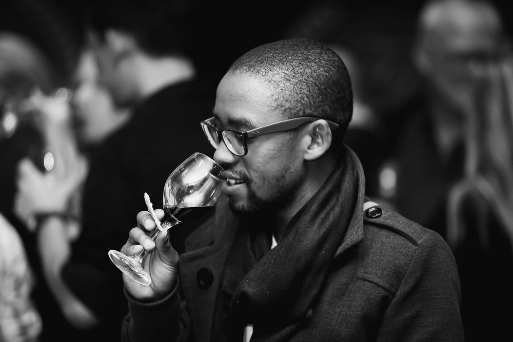 A black and white photo of a man in glasses tasting a glass of wine at a party