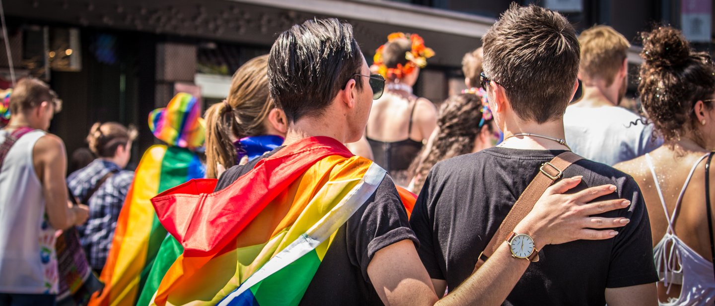 A couple with their arms around each other and a rainbow flag draped over their shoulders at a Pride parade