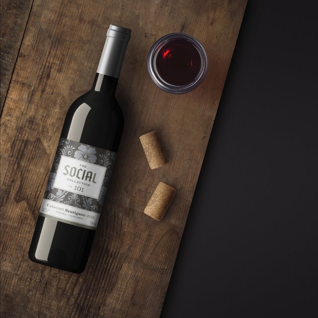 A bottle of The Social Collection Bin 101 Cabernet Sauvignon on a wooden board with a glass of red wine and corks
