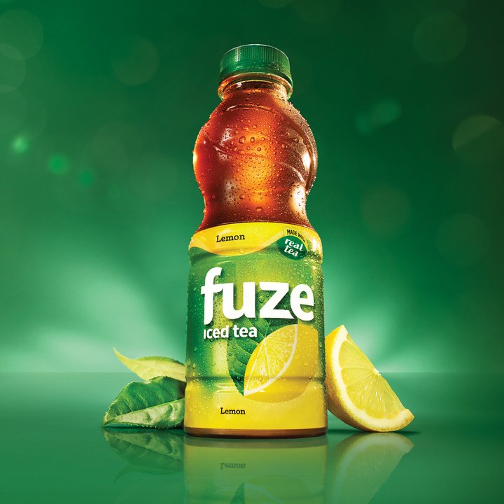 A bottle of Fuze Lemon iced tea with a lemon wedge and tea leaves, lit against a dark green background