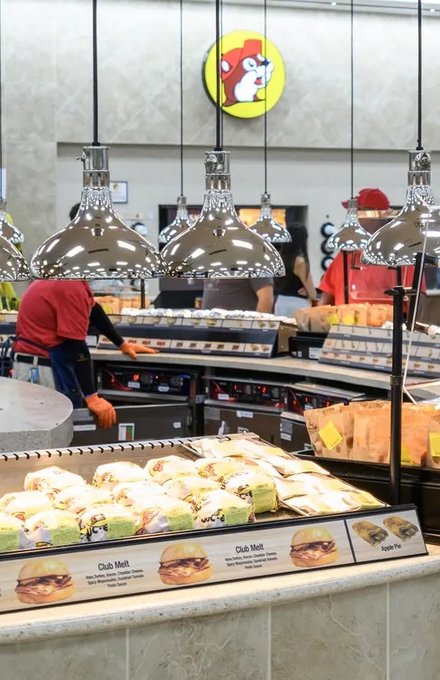 Staff in red shirts serving at the Buc-ee's food counter, with cases of prepared food under heat lamps